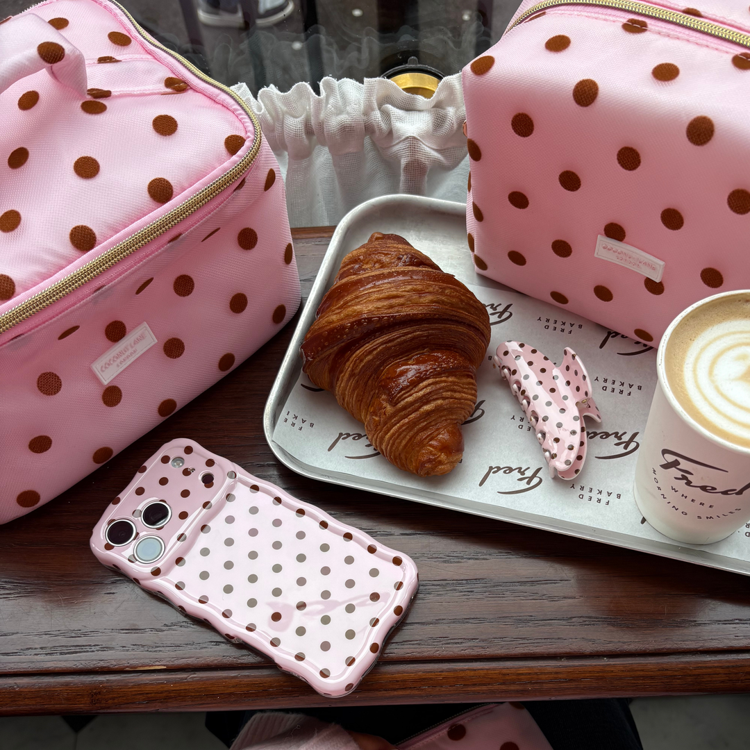 Flat lay photo in a cafe of the Dot Society pink and brown collection. The vanity case is baby pink mesh type fabric, with chocolate brown polka dots. In the photo also features the matching wavy phone case, claw clip and makeup bag.