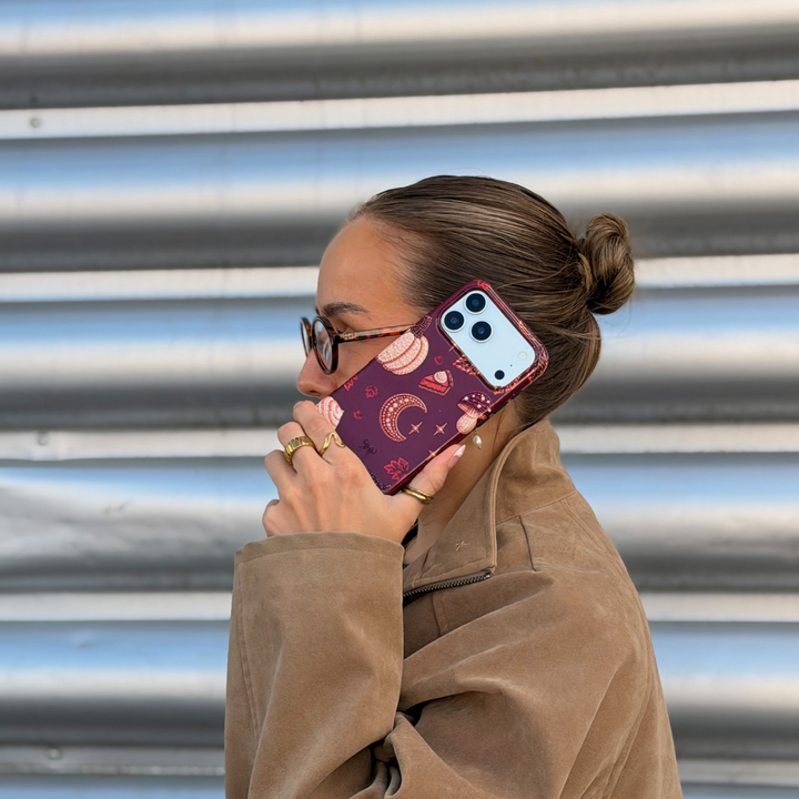 A woman with glasses and her hair in a bun holds a phone with a decorative  autumn-themed case featuring illustrations of pumpkins, a crescent moon, and a slice of pumpkin pie.