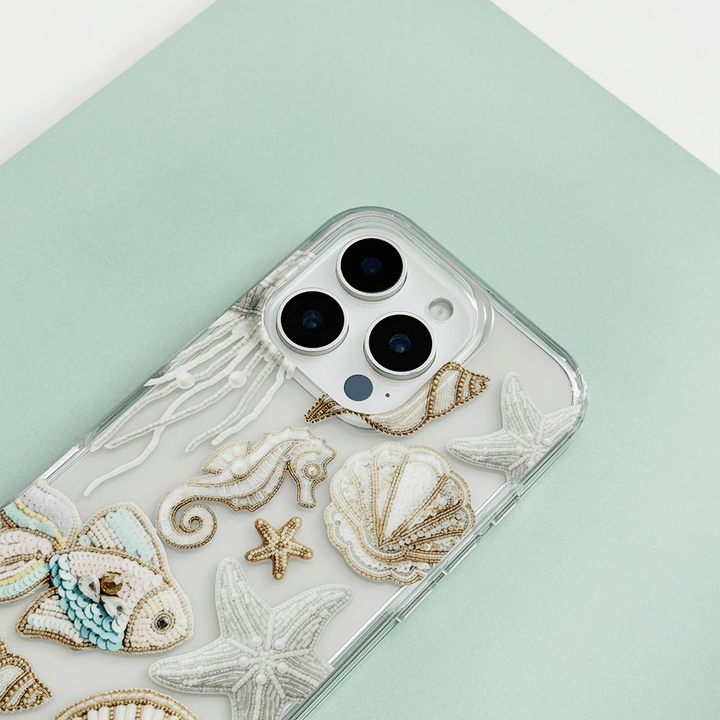 Clear phone case with embroidered shells, seahorse, starfish, and fish on a mint background.