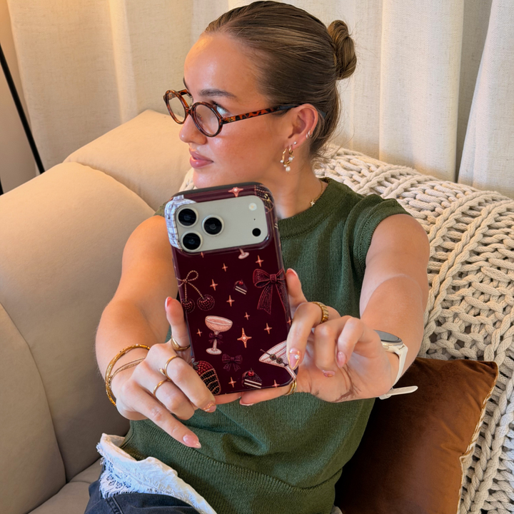 Woman in a dark green jumper with a white lace shirt underneath holds a burgundy chocolate brown phone case with decorative beaded illustrations on it, such as a bow, a strawberry, a cocktail, stars and cherries. 