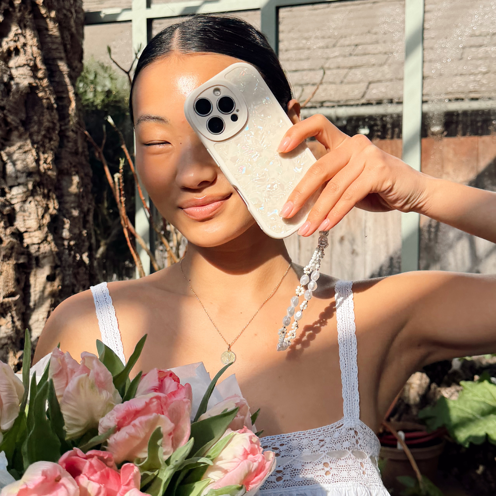 Girl in a greenhouse holding flowers posing with her Moonstone Pearl Phone Case that's shining in the sun