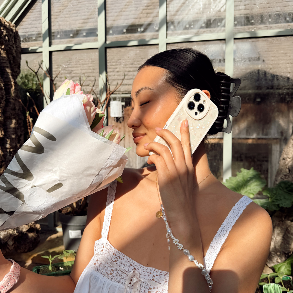 Girl in greenhouse smelling her pink flowers posing with her Moonstone Pearl Phone Case that's shining in the sun