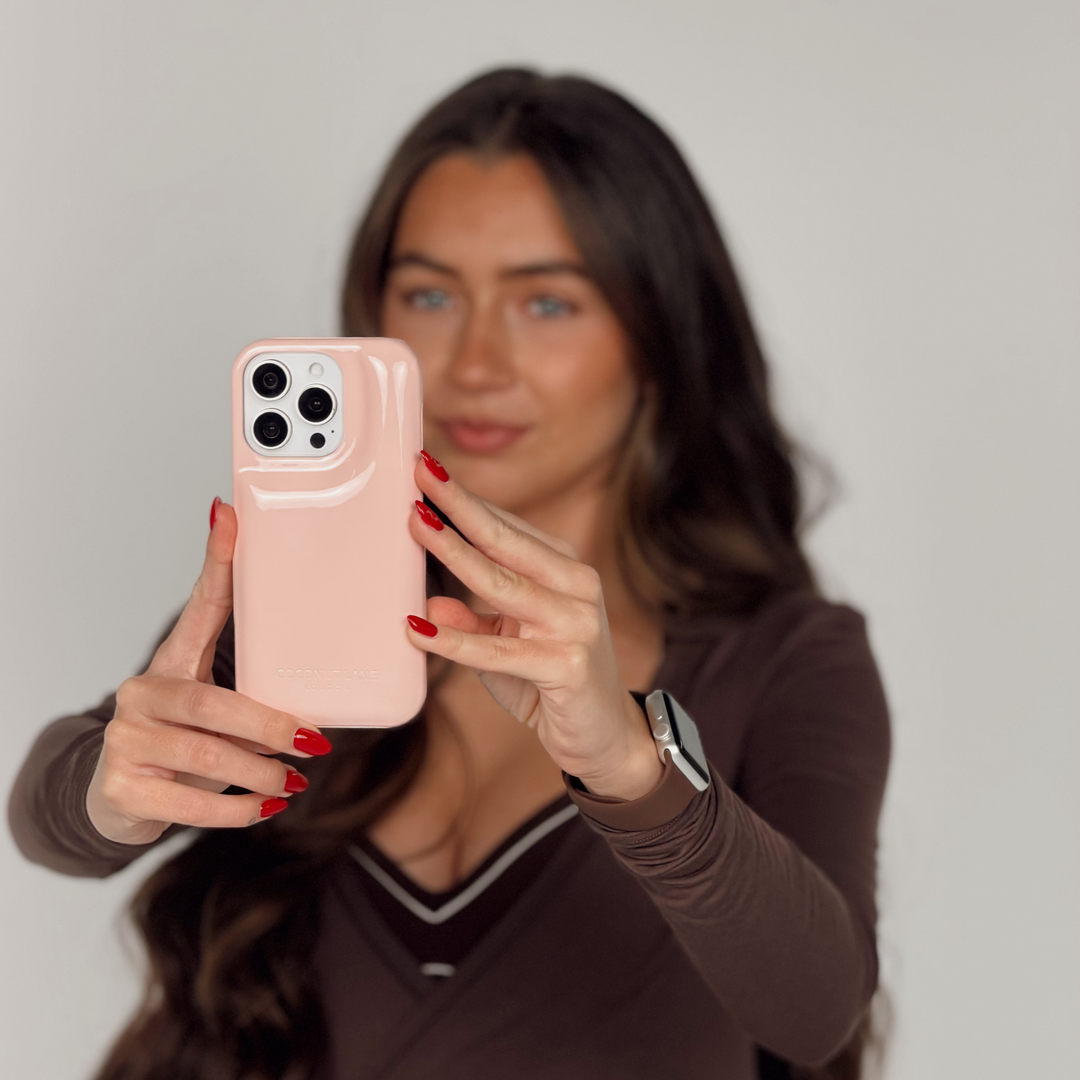 Woman in a brown pilates outfit is holding an iphone, taking a selfie. The iphone is in a glossy pastel pink Coconut Lane puffer phone case with embossed logo. On her wrist is a Coconut Lane NAKD Chocolate brown apple watch strap.