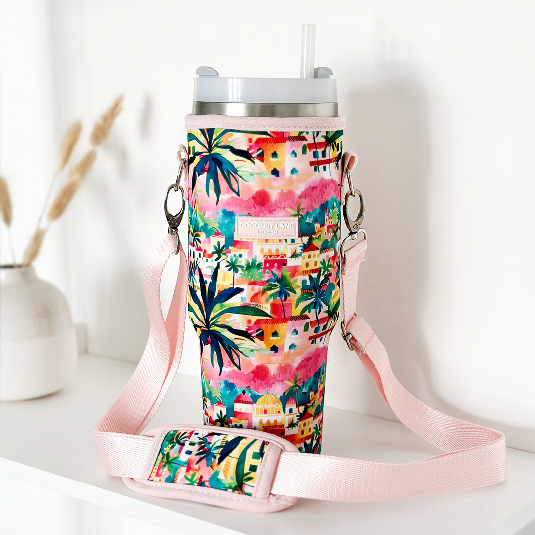 A vibrant Coconut Lane Stanley Quencher travel mug with a Portofino Dreams design, showcasing palm trees and houses in bold colors. The tumbler includes a clear lid with a straw and comes with a coordinating carry case featuring a pink strap, all set on a white surface with a small vase of dried plants in the background.