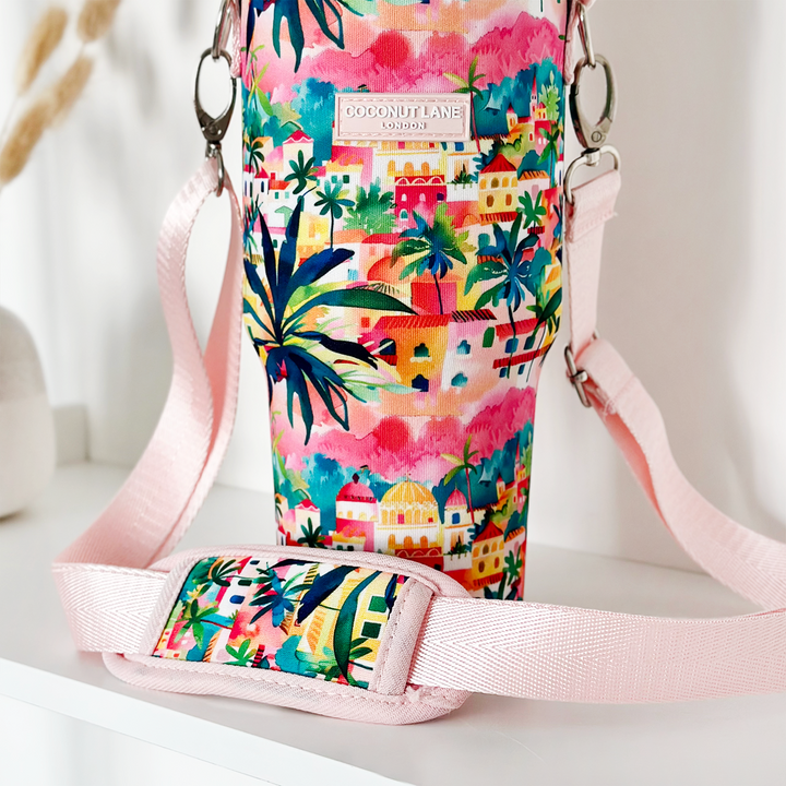 The Tumbler Carry Case - Portofino Dreams by Coconut Lane is a colorful, tropical-themed accessory adorned with vibrant illustrations of palm trees and houses. It features a pink fabric strap, silver metal clasp, and a tag that reads "Coconut Lane London." The holder is positioned on a white surface.