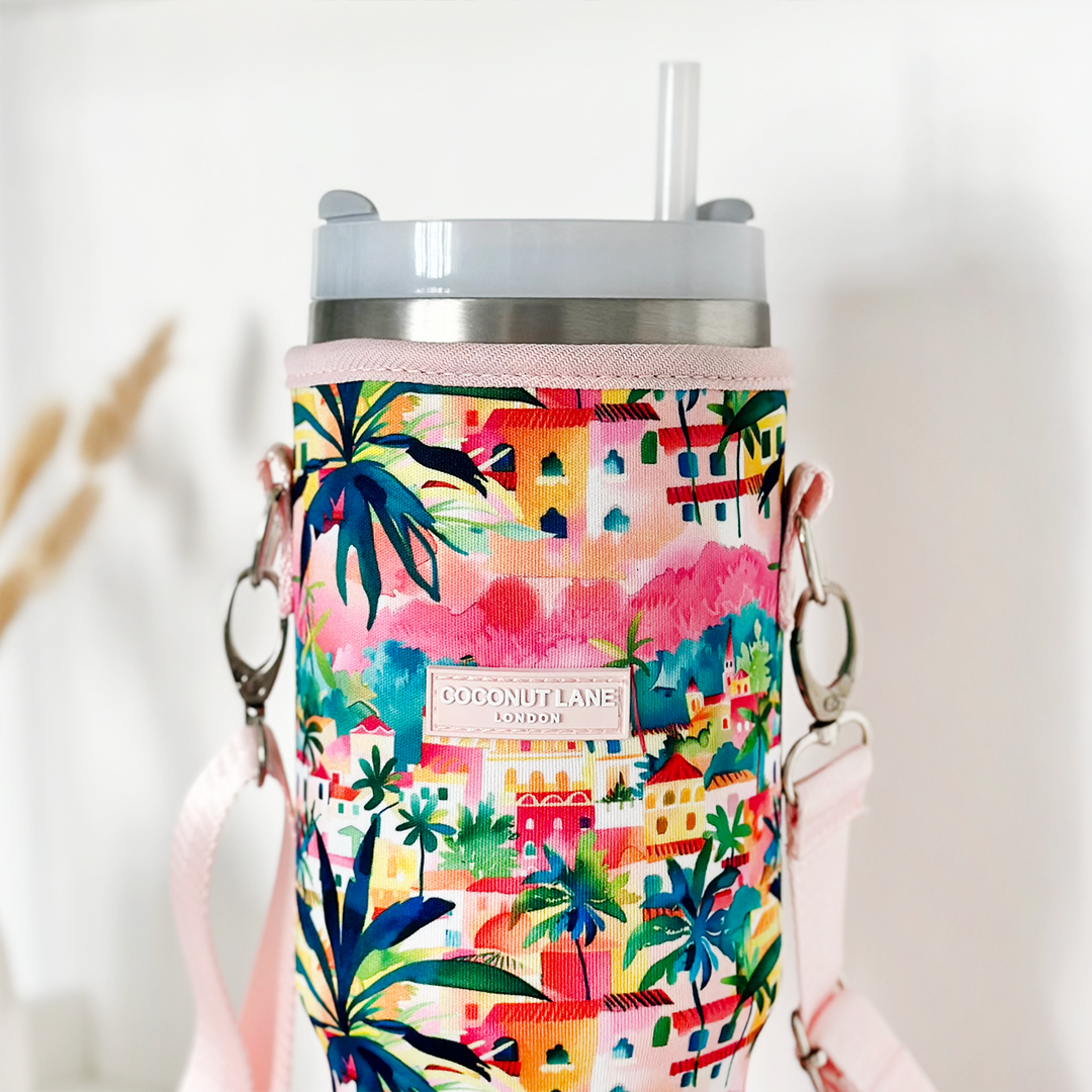 A colorful, tropical-themed insulated tumbler carry case called "Portofino Dreams" features palm trees and building prints. It has a light pink shoulder strap and silver clips. The label reads "COCONUT LANE." Inside the holder is a light gray Stanley Quencher travel mug with a straw.