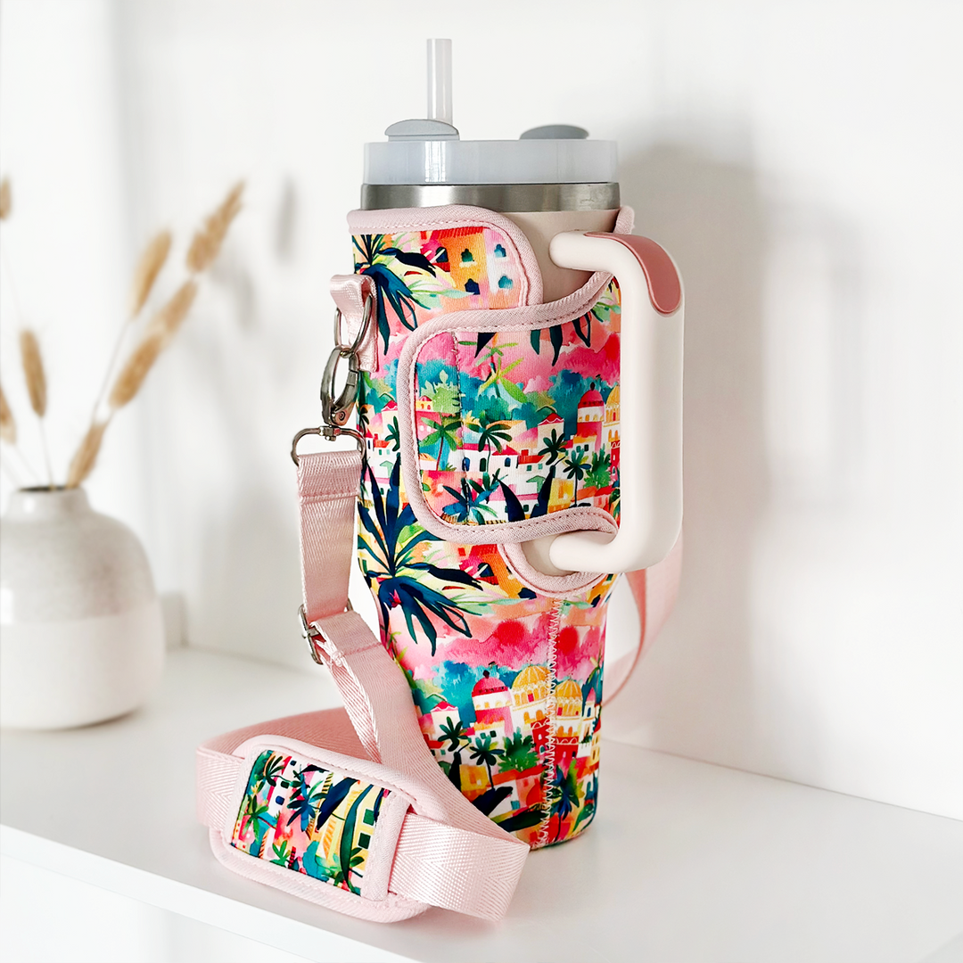 A large travel mug adorned with Coconut Lane's "Tumbler Carry Case - Portofino Dreams," featuring vibrant designs of palm trees and buildings accented by pink straps and handles, is displayed on a white surface. The case also includes an adjustable strap for easy carrying. In the background, a blurred vase with dried foliage adds a touch of elegance.