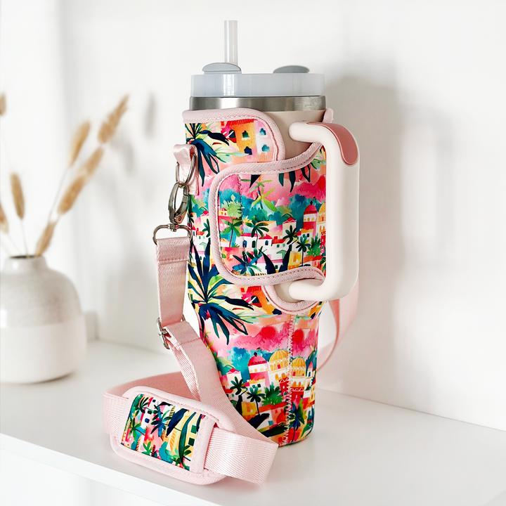 A large travel mug adorned with Coconut Lane's "Tumbler Carry Case - Portofino Dreams," featuring vibrant designs of palm trees and buildings accented by pink straps and handles, is displayed on a white surface. The case also includes an adjustable strap for easy carrying. In the background, a blurred vase with dried foliage adds a touch of elegance.