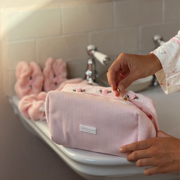 A person's hands are shown opening a light pink pointelle fabric makeup bag with a zipper. It has lace detail with flowers, set on a bathroom sink with matching pink scrunchies in the background. 