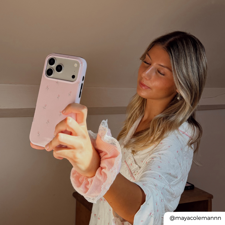 A woman wearing a white and pink floral long-sleeve top and a matching pink scrunchie holds up a light pink phone case with a subtle floral pattern.