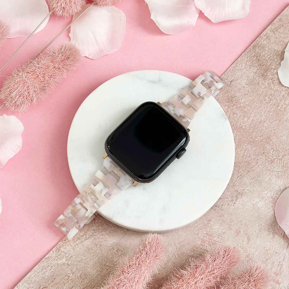 Slim Luxe Rose Apple Watch strap by Coconut Lane on a white tray, surrounded by pink flowers and petals.