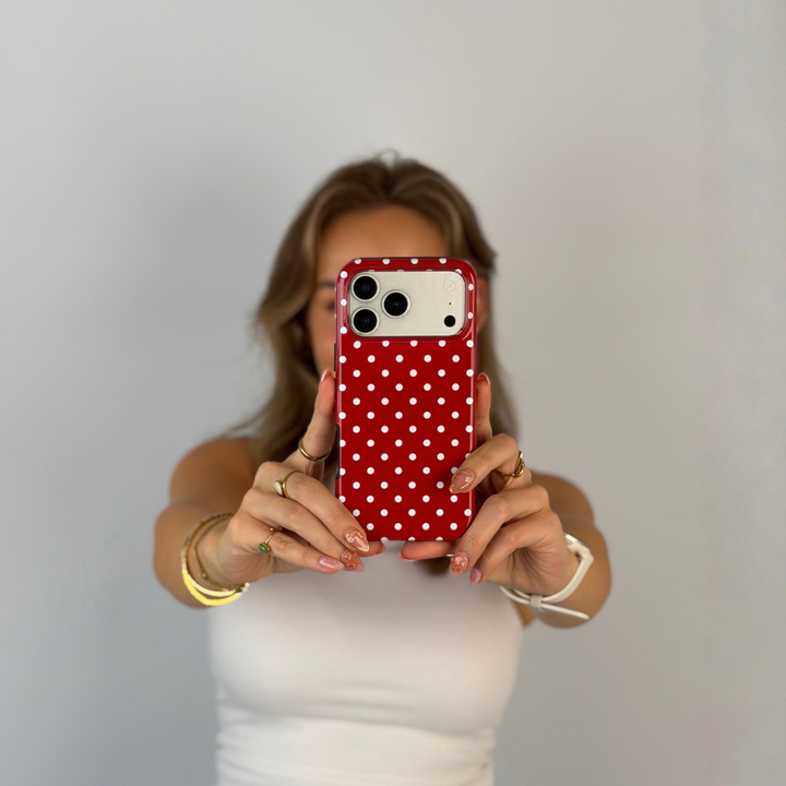 Woman holding an iphone talking a selfie. The phone is in a red and white polka dot phone case. The woman is wearing a white vest top with gold jewellery. 