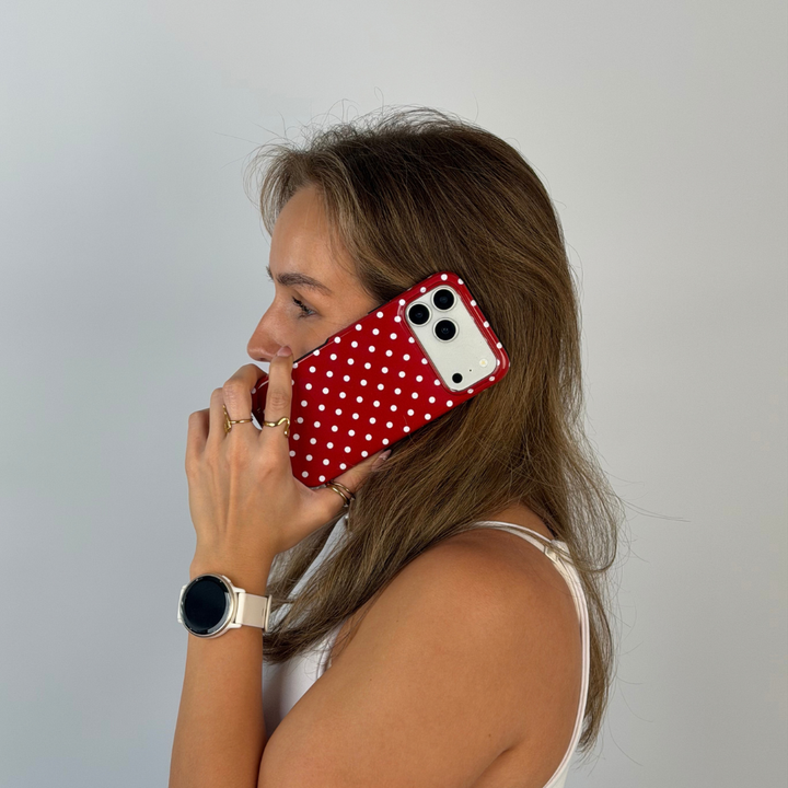 Woman holding an iphone up to her ear. The phone is in a red and white polka dot phone case. The woman is wearing a white vest top with gold jewellery. 