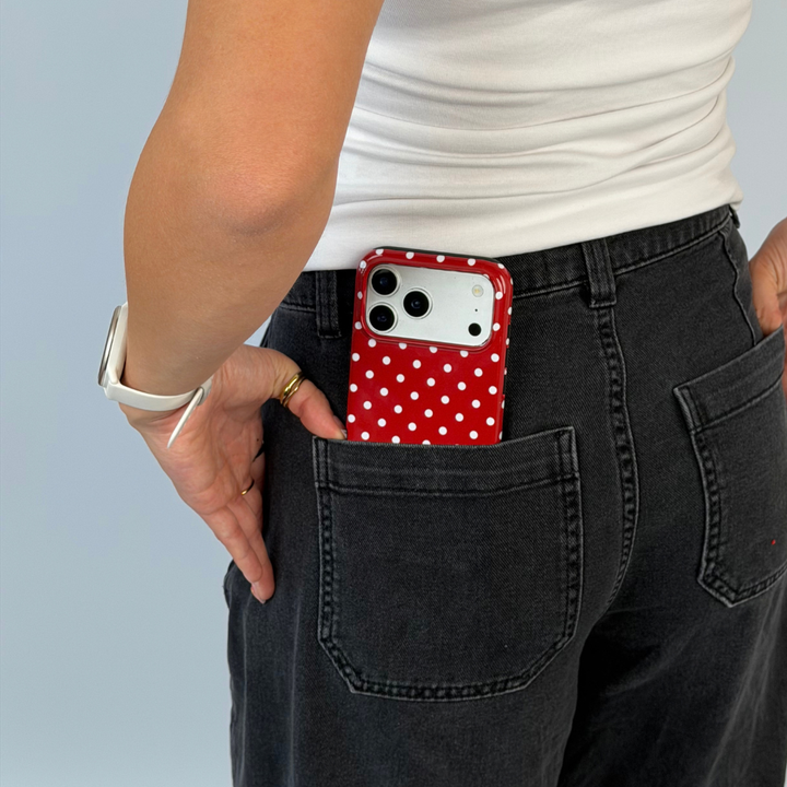 Woman with iphone in back pocket of some grey charcoal jeans. The phone is in a red and white polka dot phone case. The woman is wearing a white vest top with gold jewellery. 