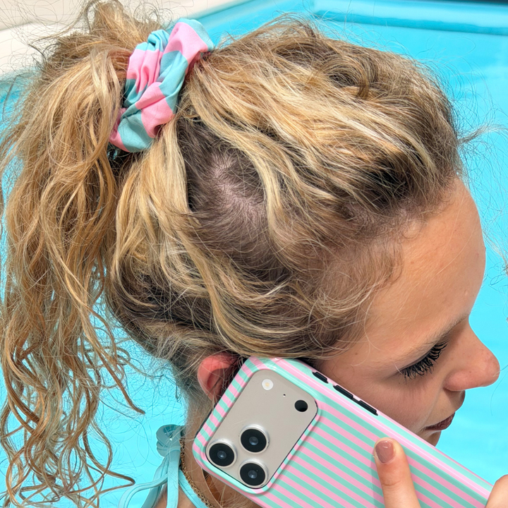 Person with a turquoise scrunchie in their hair, holding a phone with a striped case by a pool.