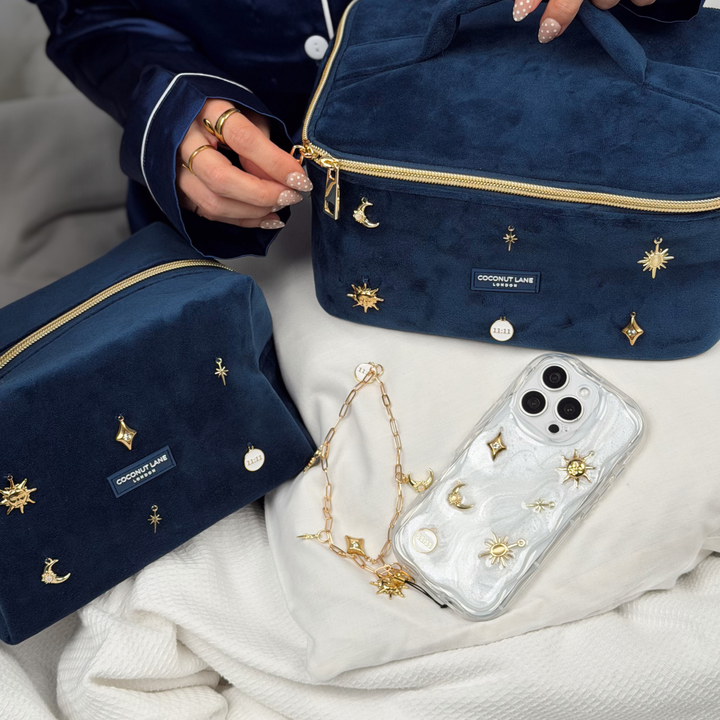 Stardust after dark charm phone case. A pearl base with luxe gold celestial charms including a moon, sun, star, diamond shape and 11:11 charm. Attached to the phone case is a matching gold chain phone strap. In the image there is a dark navy blue velvet mateiral makeup bag and vanity case,  with gold charm embellishments. 