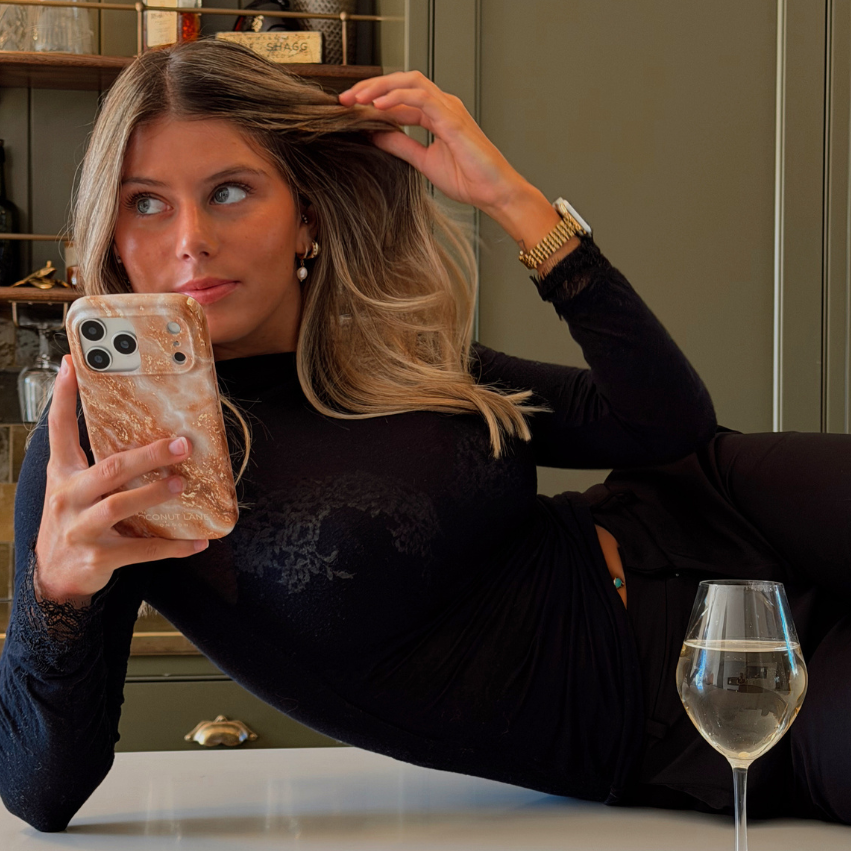 Woman with blonde hair wearing a black long sleeve top is holding an iphone in a marble effect phone case with cream, gold, light brown and white tones running through it.