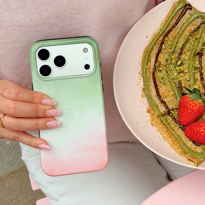 Strawberry Matcha Phone case with gradient design held by a hand with pink nails next to a dessert with strawberries and pistachio drizzle.