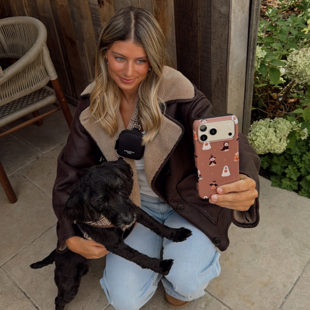 Woman in a brown aviator jacket, holding a phone case with dogs wearing spooky outfits on a brown background, whilst kneeling on the ground stroking a black dog. 