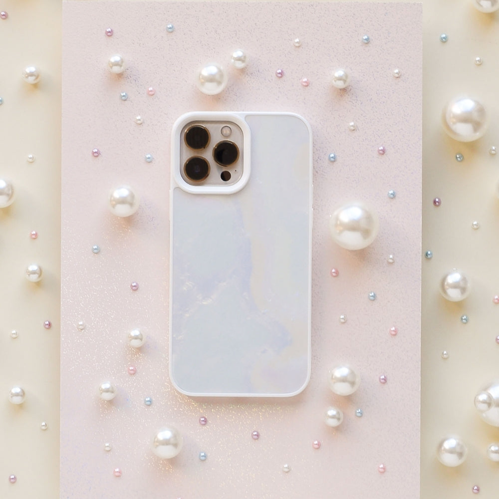 A white smartphone encased in a Coconut Lane Mother of Pearl Phone Case is centered on a light, glittery surface. The phone is surrounded by variously sized pearls scattered around, creating a delicate and elegant visual composition.