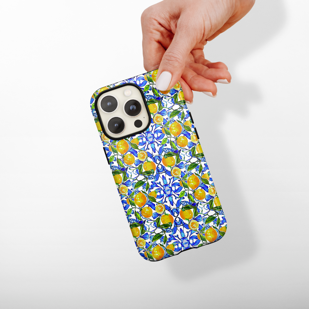 A hand holding a smartphone encased in Coconut Lane's Tough Phone Case - Amalfi Lemon. This design showcases vibrant, intricate patterns of lemons, leaves, and blue florals against a white background. It provides durable protection while ensuring the phone's camera lenses remain safely visible at the top left corner of the case.