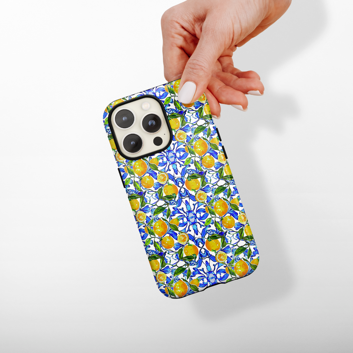A hand holding a smartphone encased in Coconut Lane's Tough Phone Case - Amalfi Lemon. This design showcases vibrant, intricate patterns of lemons, leaves, and blue florals against a white background. It provides durable protection while ensuring the phone's camera lenses remain safely visible at the top left corner of the case.