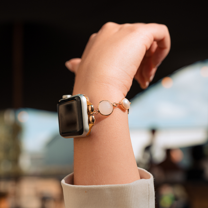 A close-up of a person's wrist showcases the Cute Stone Bracelet Apple Watch Strap by Coconut Lane, featuring a gold smartwatch with a black screen elegantly paired with the bracelet's three circular pearl-like charms. The blurred background depicts an outdoor setting with a light blue sky and some greenery.