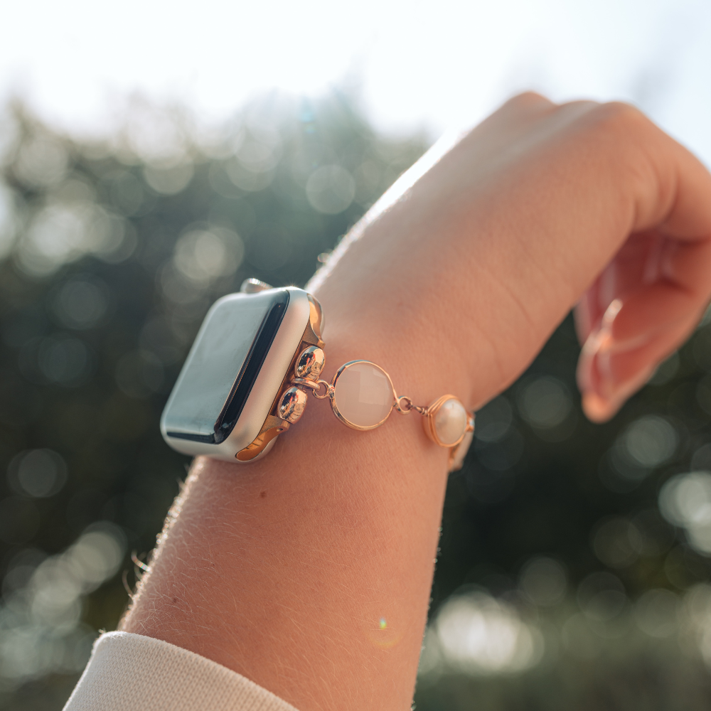 A close-up of a person's wrist against a blurred outdoor background. The wrist is adorned with Coconut Lane's Cute Stone Bracelet Apple Watch Strap, featuring three circular charms.