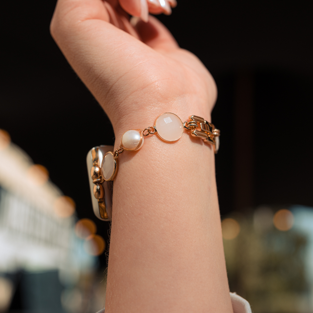 A close-up photo of a person's wrist adorned with a delicate gold Apple Watch strap. The Cute Stone Bracelet Apple Watch Strap by Coconut Lane features circular white and pearl-like charms. The background is blurred, with warm lighting suggesting an indoor setting. The person's hand is slightly raised.