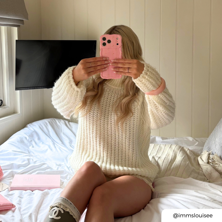 Girl with blonde hair sat in a shepherds hut, holding a wavy pink striped phone case with small spicy margarita illustrations.
