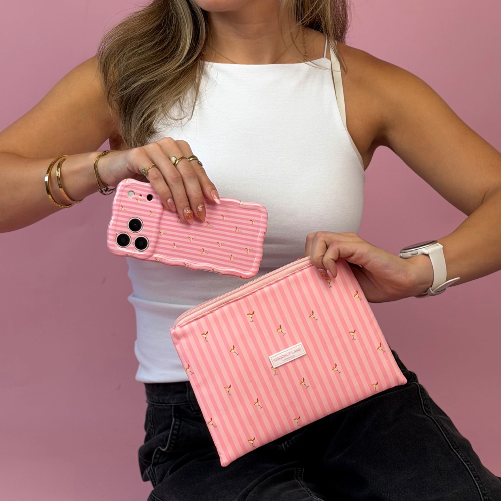A woman holds a pink striped phone case in one hand and a pink zippered cosmetic pouch with vertical pink stripes and small spicy margarita illustration prints on it. 