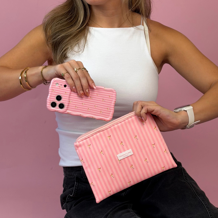 A woman holds a pink striped phone case in one hand and a pink zippered cosmetic pouch with vertical pink stripes and small spicy margarita illustration prints on it. 