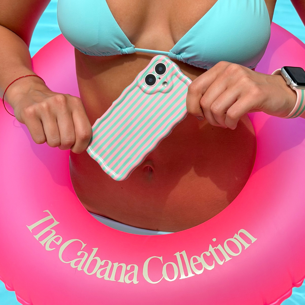 Girl in turquoise bikini holding a wavy phone case on a pink inflatable ring with 'The Cabana Collection' branding.