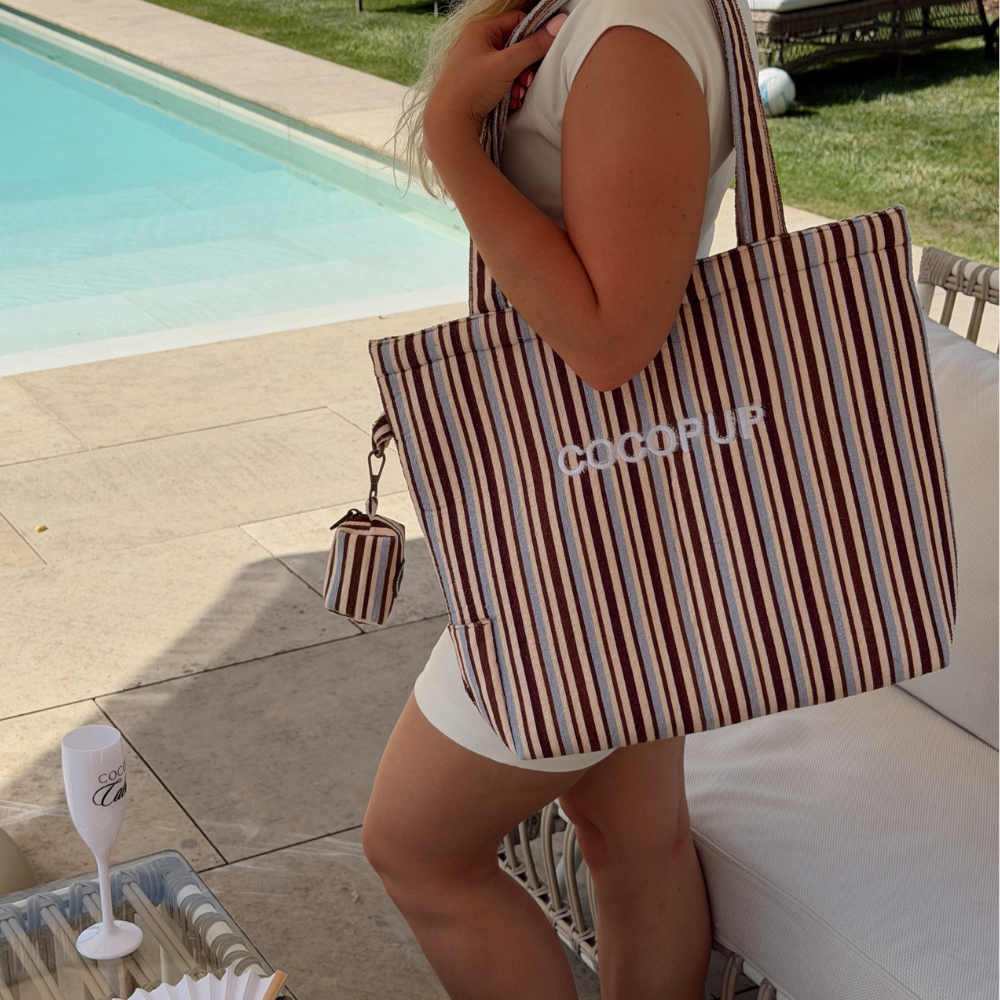 Woman poolside with a brown and blue striped tote bag, wearing white; drink set nearby.
