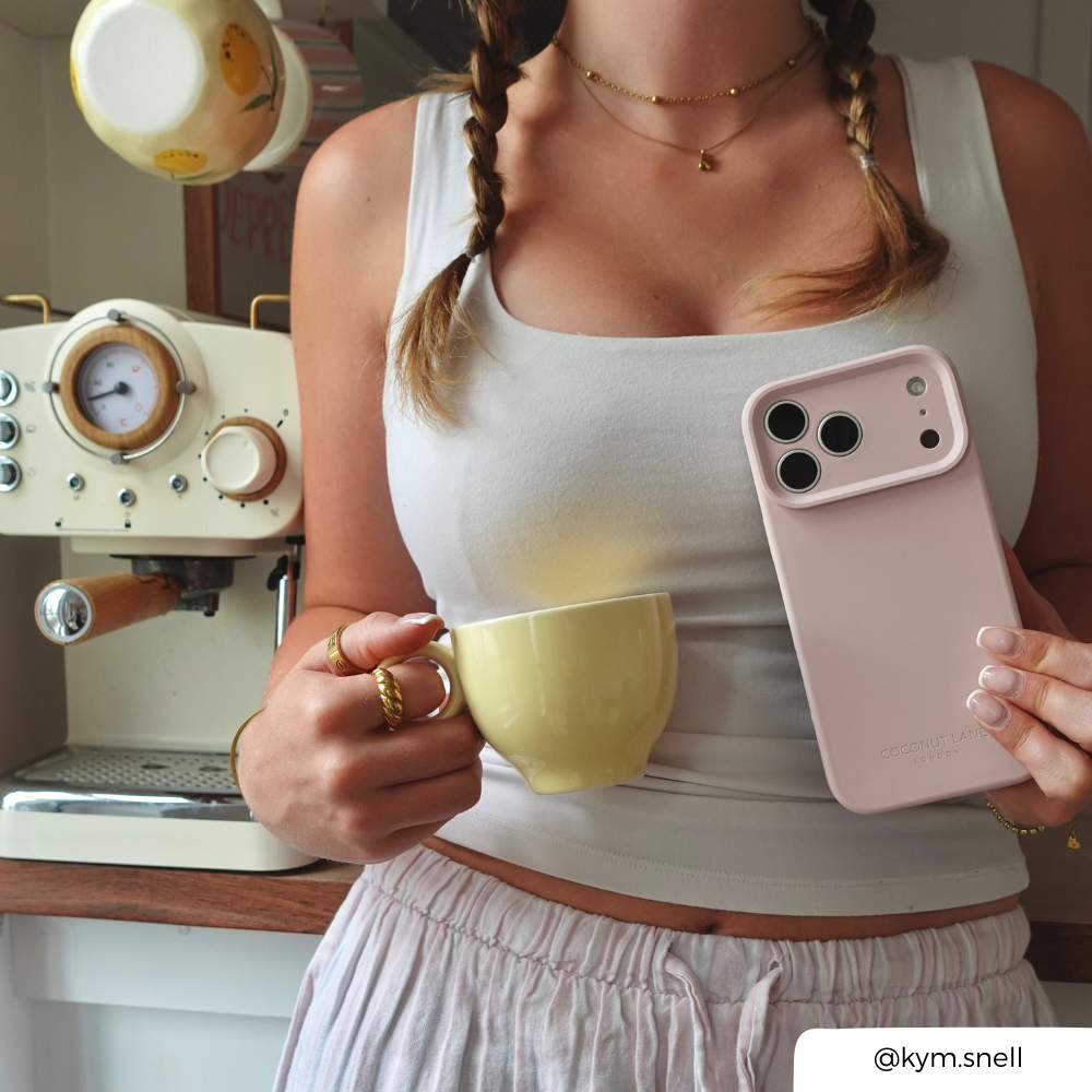 a women holding a coffee in a soft, pastel kitchen setting, holding an iphone in a pastel pink phone case. 