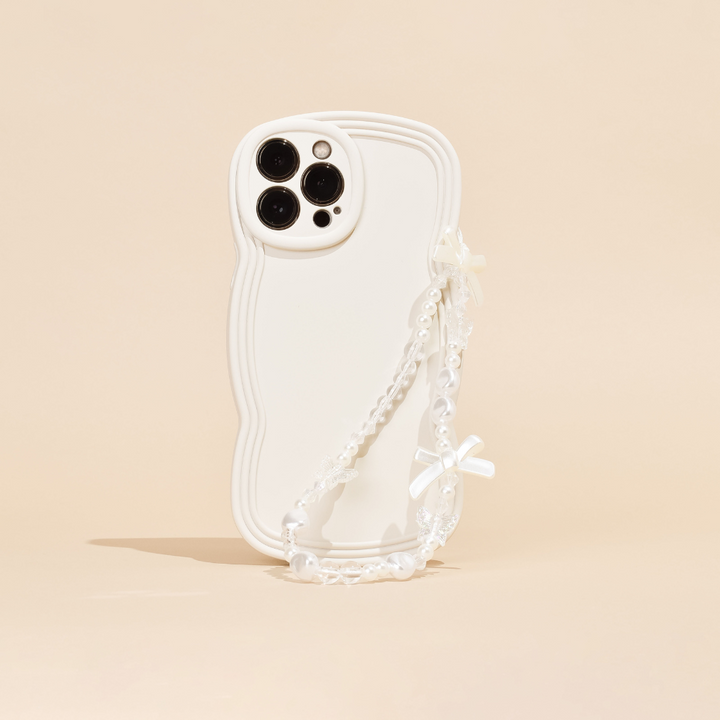 A white phone case with a wavy edge design is pictured against a beige background. This chic phone accessory from Coconut Lane features three camera cutouts and the Beaded Phone Strap - Bows & Butterflies, a decorative pearl chain with small white bows and butterflies attached, hanging from one side. The beaded phone strap adds an extra touch of elegance.