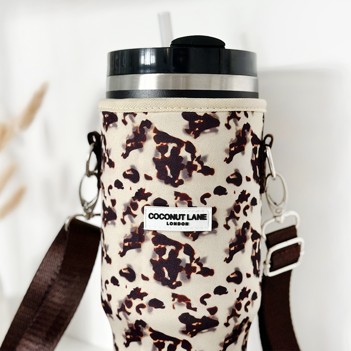 A close-up shot of a stylish travel mug with a straw in an Ivory Tort Tumbler Carry Case. The case, which doubles as a sleeve, has an attached strap and features a label that reads "Coconut Lane." The background is white and blurred, focusing attention on the vibrant print and texture.