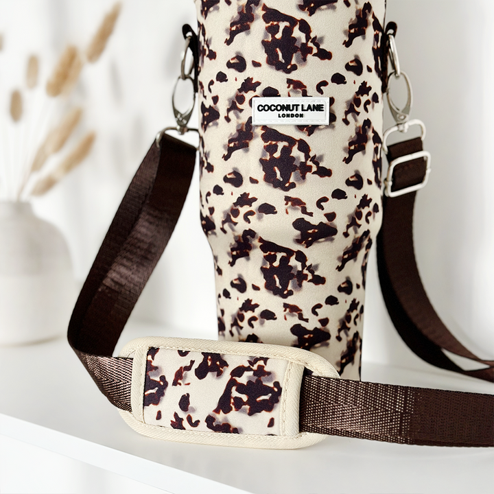 A close-up of the stylish Tumbler Carry Case - Ivory Tort, featuring an elegant animal-print design and a brown adjustable strap, perfect for carrying your travel mug. The brand label on the case reads "Coconut Lane." In the background, a blurred white vase with dried pampas grass is visible on a white surface.