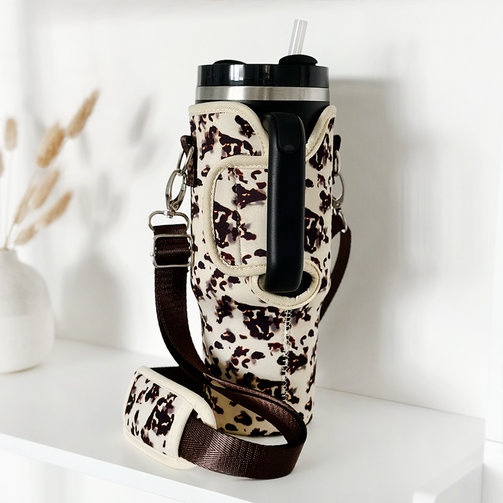 A large Stanley Quencher tumbler with a black lid and straw is encased in Coconut Lane's Tumbler Carry Case - Ivory Tort. The holder features a black shoulder strap and is placed on a white shelf. In the background, there is a white vase with dried grasses.