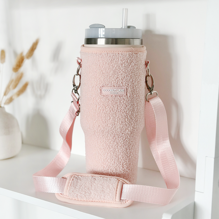 A stainless steel Stanley Quencher 40oz travel mug, adorned with a Coconut Lane Tumbler Carry Case - Teddy Love-A-Lot featuring handles and a shoulder strap, is placed on a white shelf. In the background, there are a few dried plants in a white vase.