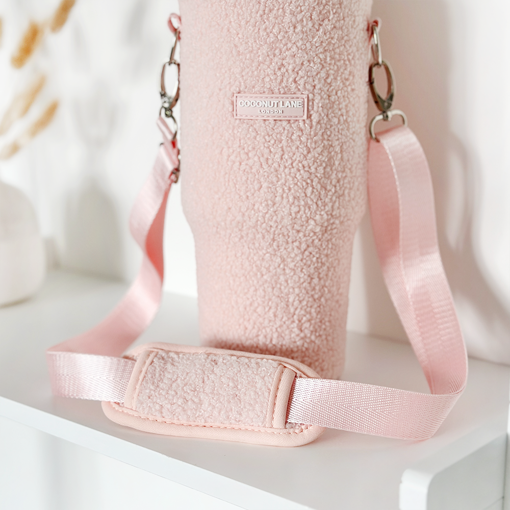 A Tumbler Carry Case - Teddy Love-A-Lot from Coconut Lane is displayed on a white background. This textured pink holder features metallic clasps, an adjustable strap with a cushion pad for shoulder comfort, and a rectangular label that reads "COCONUT LANE." It's ideal for holding a Stanley Quencher 40oz travel mug.