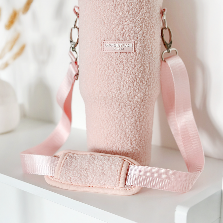 A Tumbler Carry Case - Teddy Love-A-Lot from Coconut Lane is displayed on a white background. This textured pink holder features metallic clasps, an adjustable strap with a cushion pad for shoulder comfort, and a rectangular label that reads "COCONUT LANE." It's ideal for holding a Stanley Quencher 40oz travel mug.