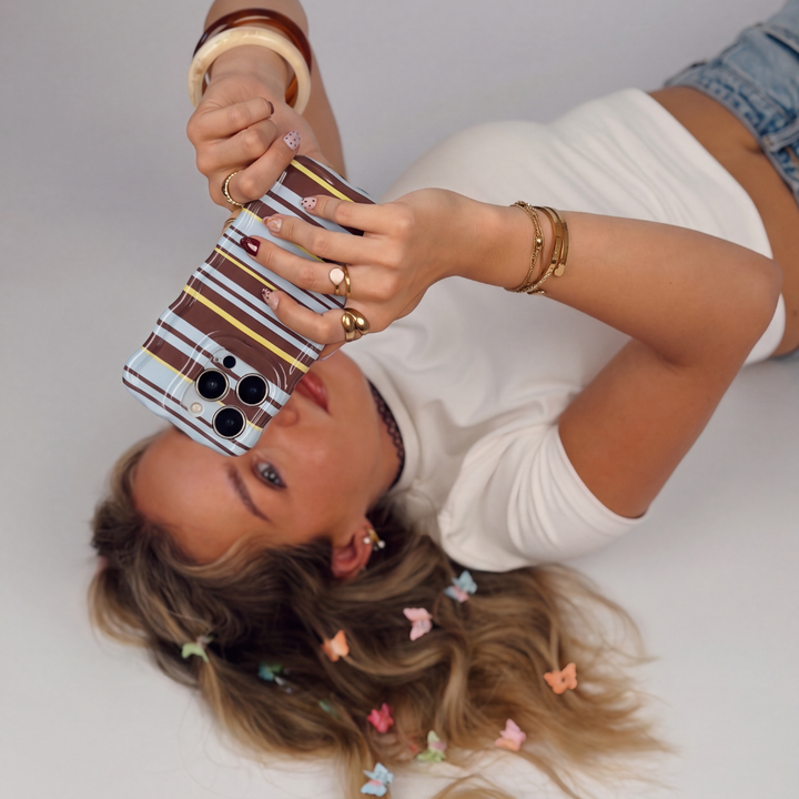 girl on the floor holding a wavy brown, blue and yellow phone case. Wearing a white t-shirt and jeans