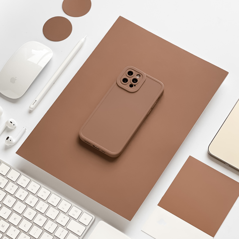 A brown smartphone with a matching NAKD Phone Case - Chocolate Brown from Coconut Lane is centered on a brown rectangular mat. Surrounding the mat are various items: a white computer mouse, white earbuds, a stylus, a keyboard, circular brown coasters, a notebook, and color swatch samples.