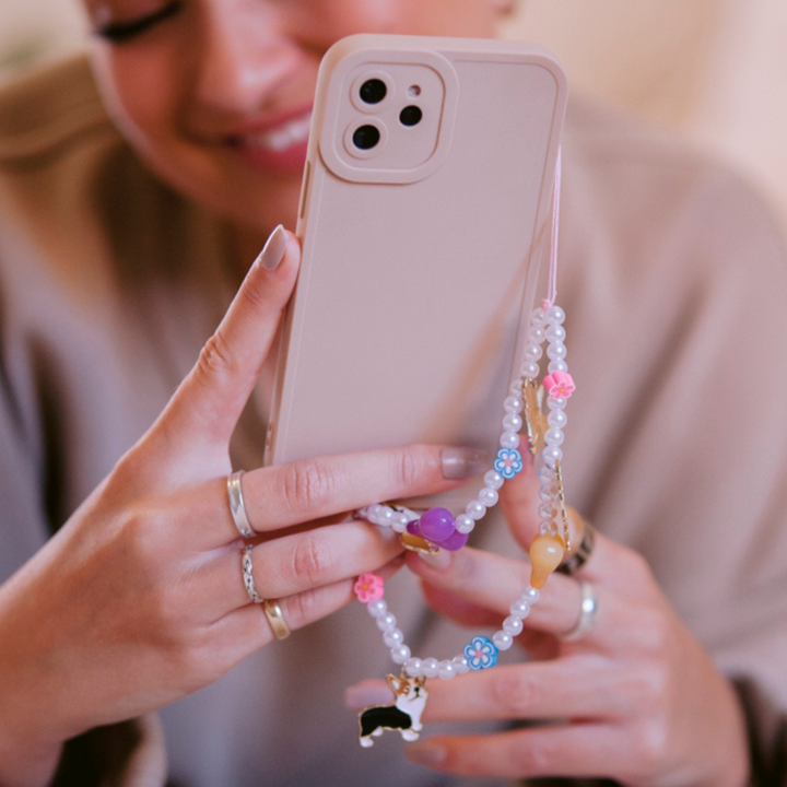 A person is holding a NAKD Phone Case - Nude by Coconut Lane, featuring a soft flexible TPU material in pink, adorned with a colorful beaded strap with various charms, including one shaped like a cat. They are wearing multiple rings on their fingers and have a partially visible face showing a slight smile.