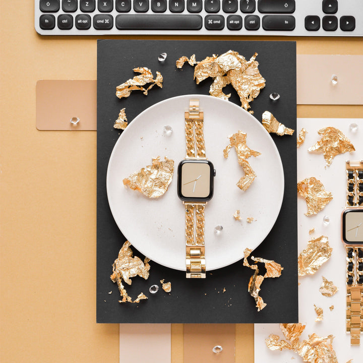 A stylish Dual-Chain Apple Watch Strap by Coconut Lane with a metallic band is placed on a white round plate. Surrounding the plate are scattered pieces of gold foil and tiny decorative gems. The backdrop includes parts of a computer keyboard at the top, set against a black and tan background, highlighting its Zinc Alloy Adjustable strap.