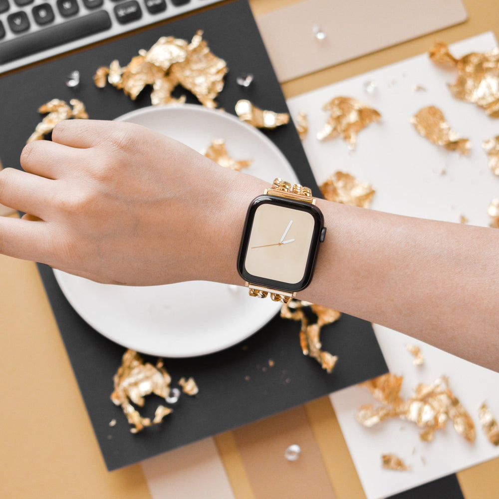 A hand wearing a stylish smartwatch with a Coconut Lane Dual-Chain Apple Watch Strap and a beige watch face is shown in close-up. The background features a white plate, a keyboard, and scattered gold foil pieces on a black and beige surface, creating a luxurious aesthetic.