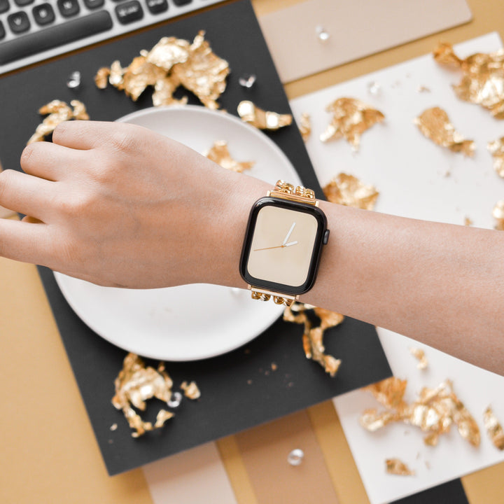 A hand wearing a stylish smartwatch with a Coconut Lane Dual-Chain Apple Watch Strap and a beige watch face is shown in close-up. The background features a white plate, a keyboard, and scattered gold foil pieces on a black and beige surface, creating a luxurious aesthetic.