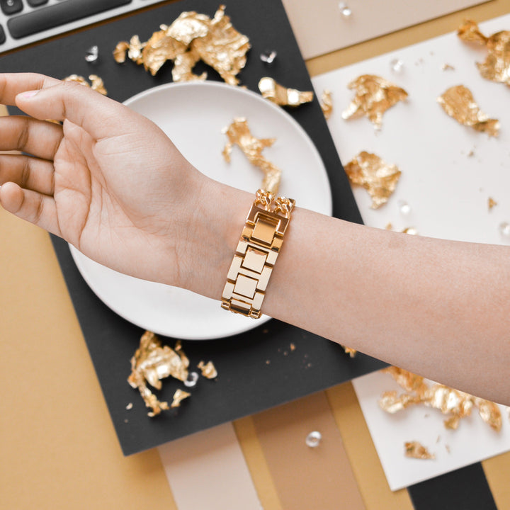 A hand adorned with Coconut Lane's Dual-Chain Apple Watch Strap is extended over a striking background of black, gold, and beige paper. Shiny, irregular gold foil pieces are scattered around, along with a partial view of a white plate and a keyboard in the background. The dual-chain design of the Apple Watch strap complements the elegant setup perfectly.