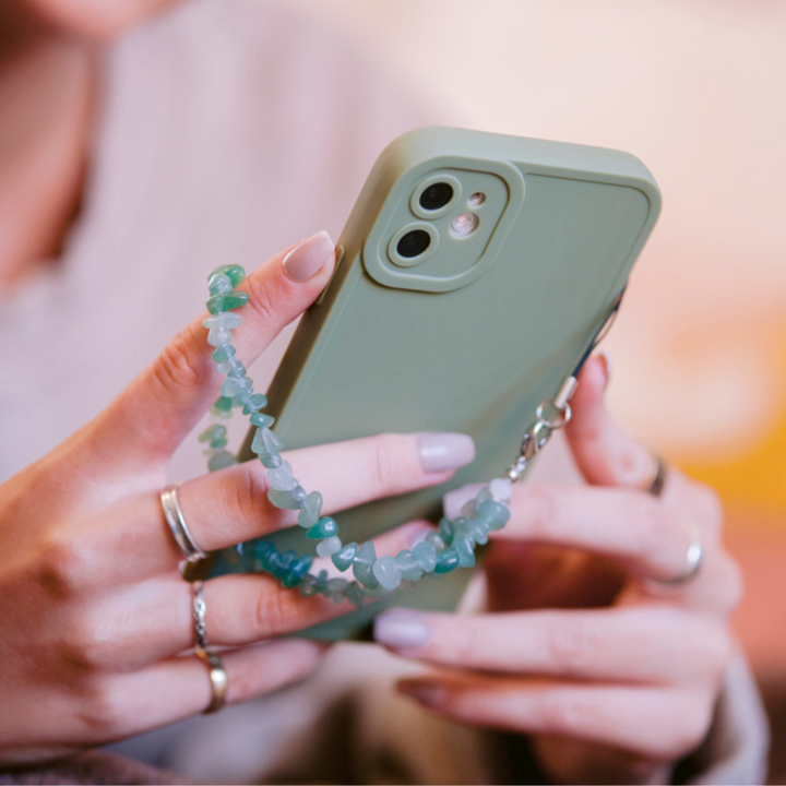 A person holds a NAKD Phone Case - Sage from Coconut Lane, complemented by a matching beaded phone charm. The phone is encased in soft, flexible TPU that ensures full camera protection. The person’s nails are painted light pink, and they are wearing multiple rings on their fingers. The softly blurred background accentuates the phone and accessories.